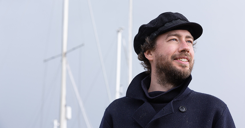 Historian Oskar Cox Jensen in a coat and hat smiling with boat masts in the background Historian Oskar Cox Jensen in a coat and hat smiling with boat masts in the background
